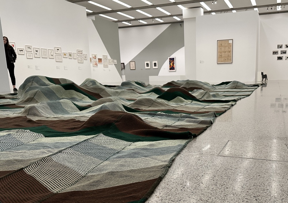 Ein Teppich, der am Boden im Museum liegt. Hügel bauen sich darunter auf. Wahrscheinlich Stofftiere. Das Werk stammt von Mike Kelley
